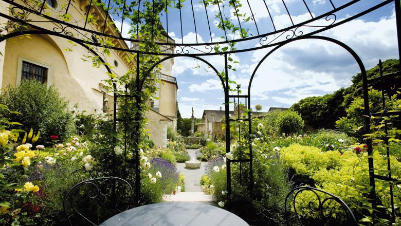 Ein idyllischer Garten mit Rosen und einem schmiedeeisernen Pavillon vor einem gelben Geb&auml;ude.