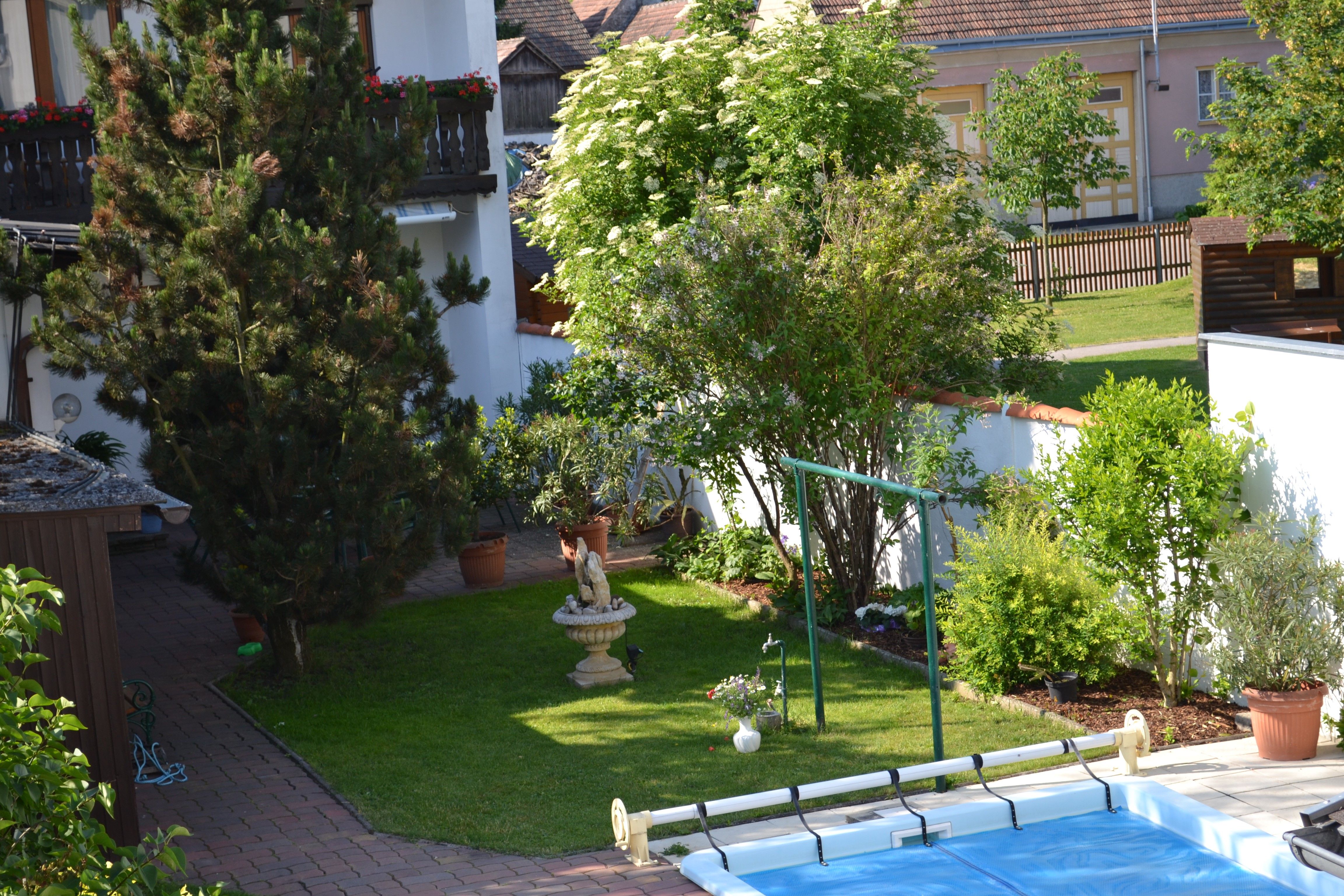 Ein gepflegter Garten mit Bäumen, Sträuchern, einer Statue und einem Pool. Im Hintergrund sind ein Haus und ein Zaun zu sehen.