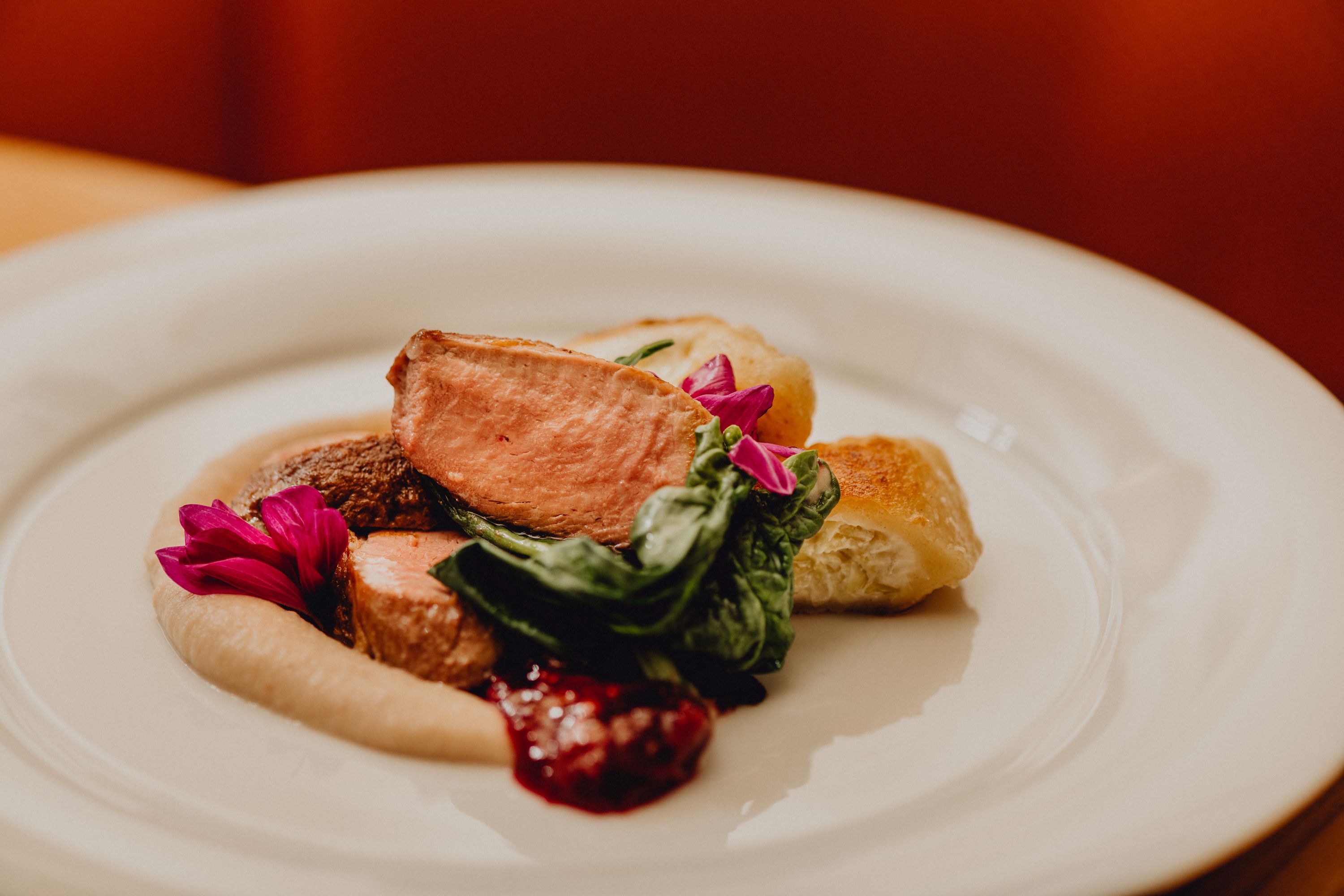 Ein Teller mit rosa gebratener Ente, Rahmtascherl, Maroni-Püree und grünen Blättern, garniert mit essbaren Blüten.
