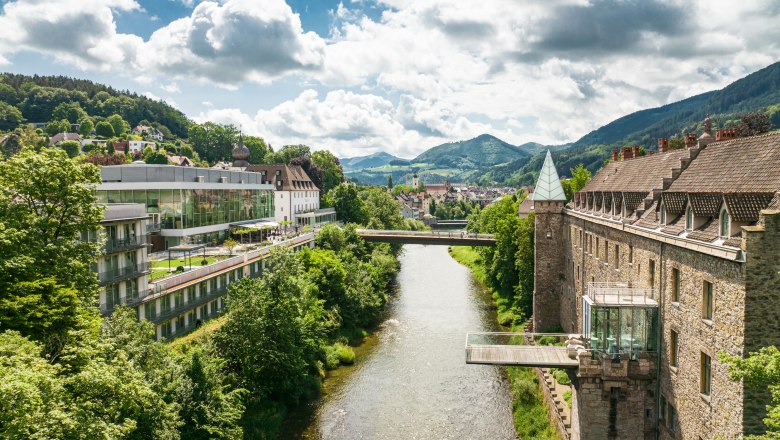 Das Schloss an der Eisenstrasse, © Dominik Stixenberger Panorama einer Flusslandschaft mit Schloss und moderner Architektur, umgeben von grünen Hügeln.