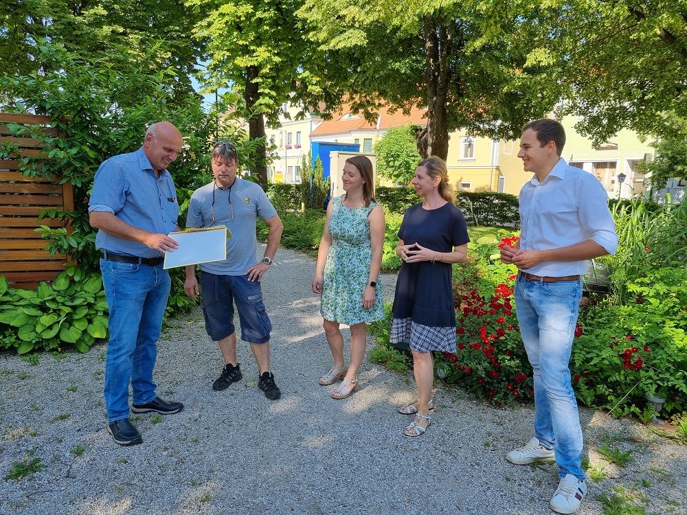 Fünf Personen stehen auf einem Kiesweg in einem Garten und unterhalten sich. Einer hält ein Dokument in der Hand.