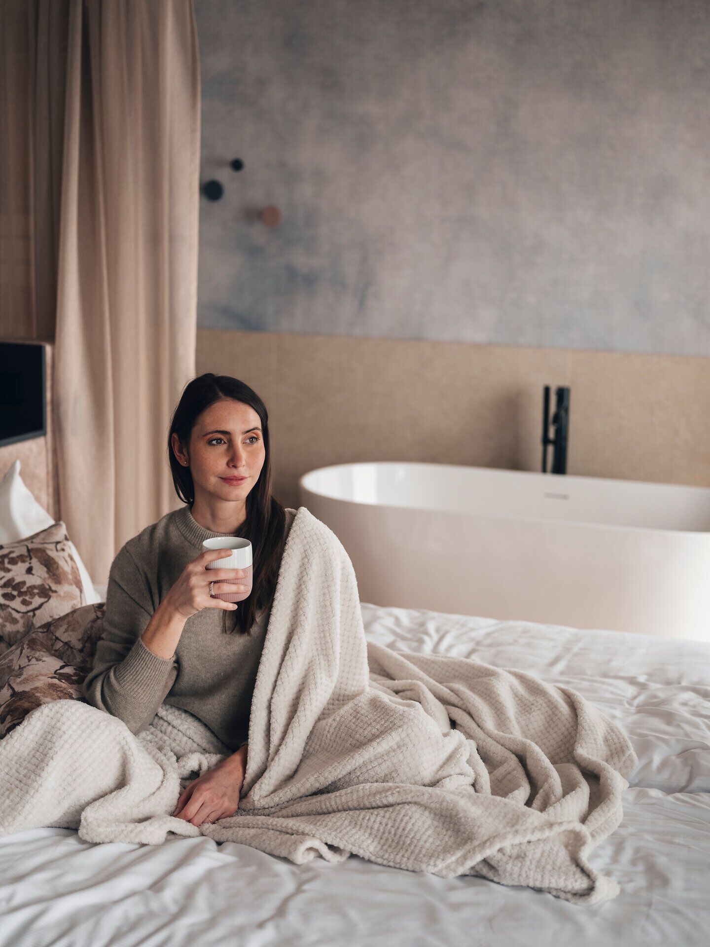 In einem stilvollen Hotelzimmer, umgeben von sanften Erdtönen, genießt eine Frau eine Tasse Kaffee und lässt den Blick über die ruhige Landschaft schweifen. Die gemütliche Atmosphäre und die weichen Textilien laden zum Entspannen ein, während die sanften Lichtverhältnisse eine harmonische Stimmung schaffen.
