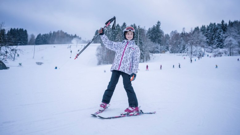Ein Kind in Skiausr&uuml;stung steht auf einer verschneiten Piste und h&auml;lt Skist&ouml;cke in die Luft. Im Hintergrund sind B&auml;ume und andere Skifahrer zu sehen.