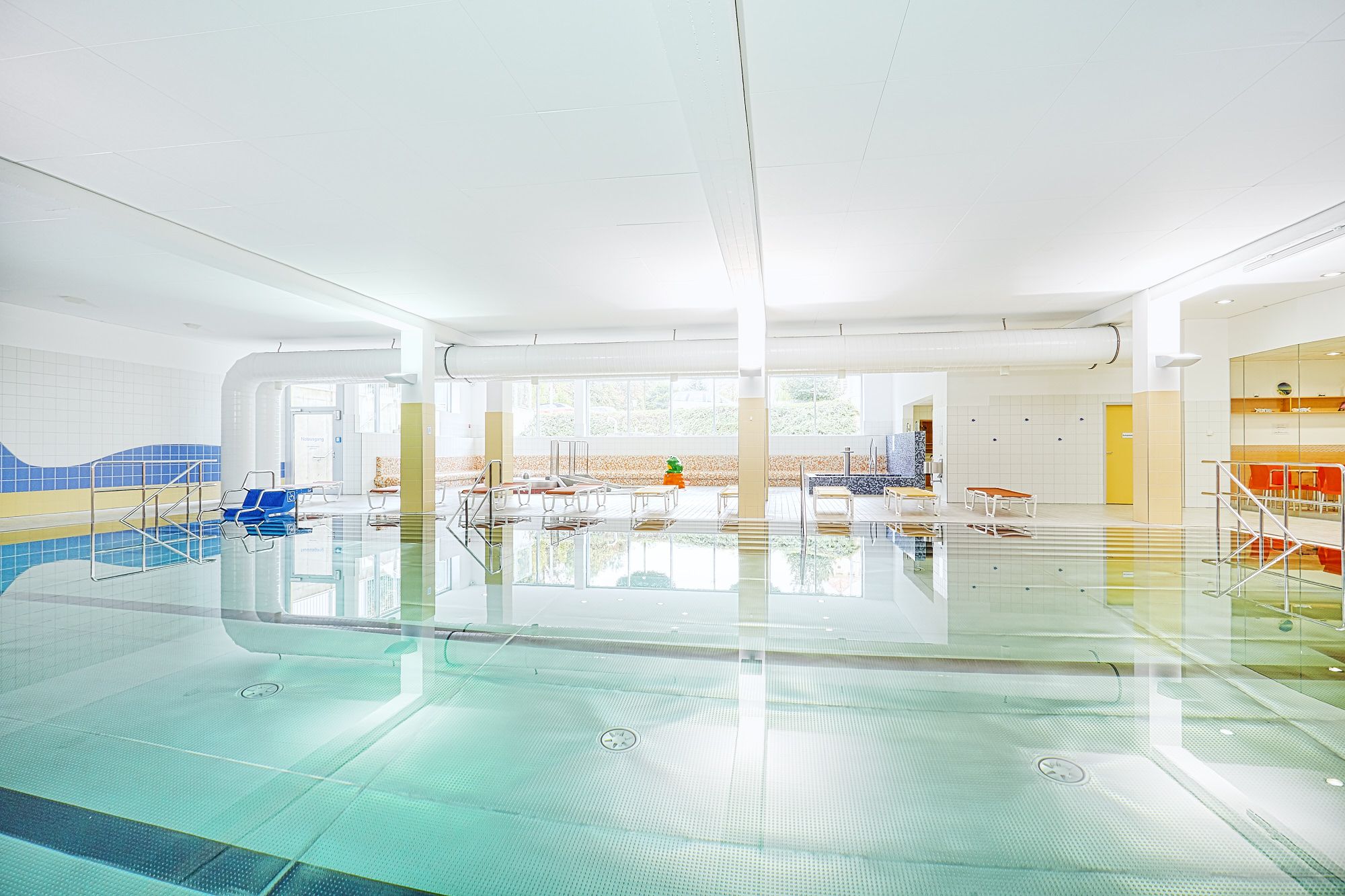 Innenansicht eines hellen Hallenbads mit Schwimmbecken, Liegestühlen und großen Fenstern.