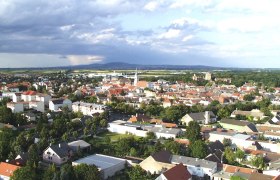Luftaufnahme von Bruck an der Leitha mit Häusern und Kirche, umgeben von grüner Landschaft und bewölktem Himmel.