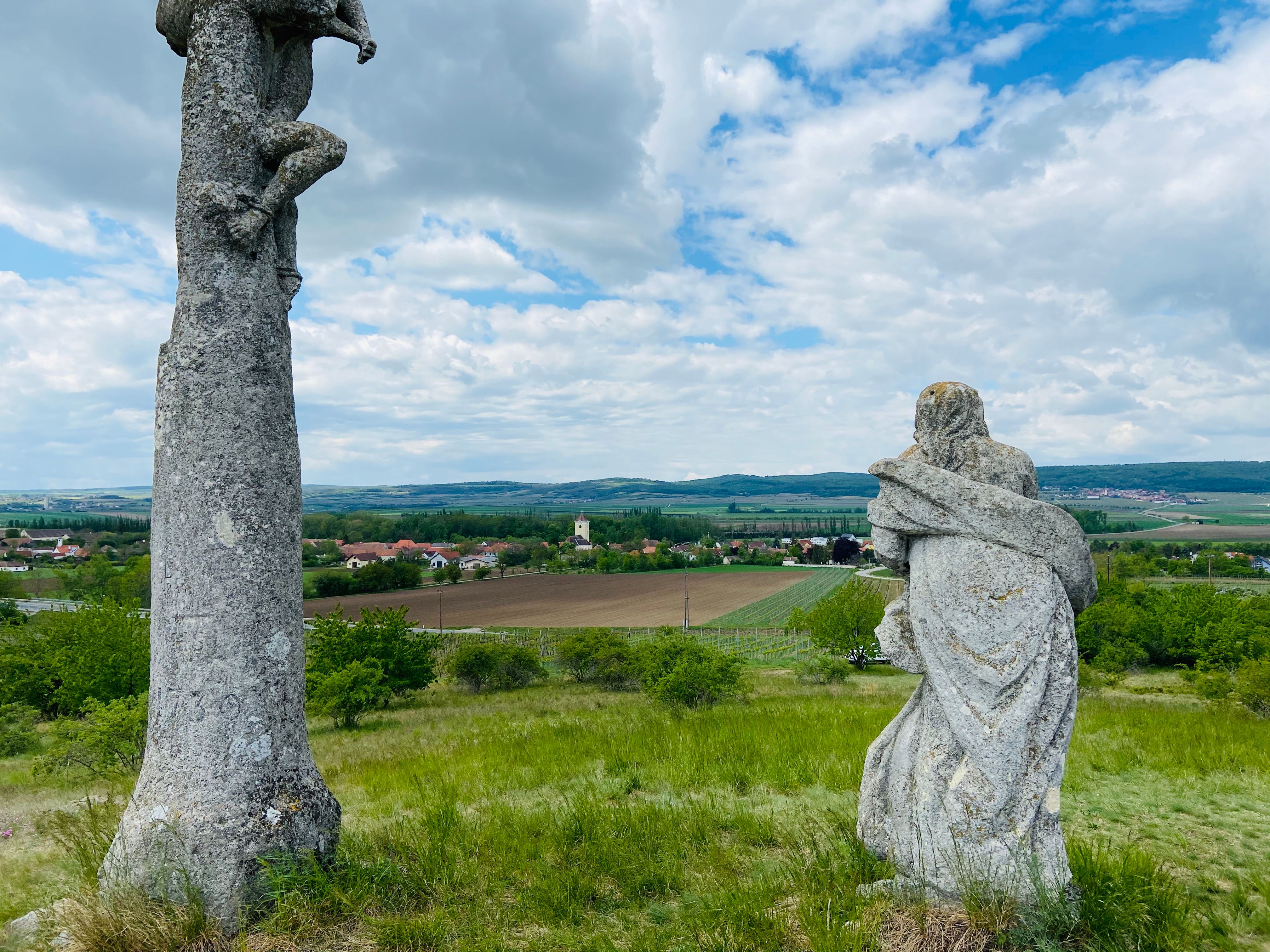 Zwei Steinskulpturen auf einem Hügel mit Blick auf eine ländliche Landschaft und ein Dorf im Hintergrund.