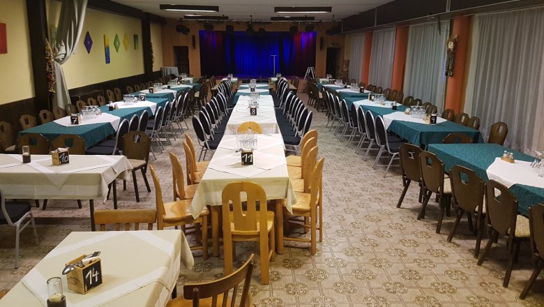 Ein leerer Veranstaltungssaal mit gedeckten Tischen und einer Bühne im Hintergrund.