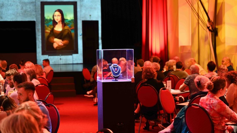 Ein Theater mit Menschen an Tischen, ein beleuchtetes Schmuckst&uuml;ck in einer Vitrine und ein Bild der Mona Lisa im Hintergrund.