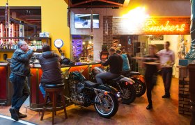 Bar mit Motorrädern als Sitzgelegenheiten, Neonlicht und Gästen.