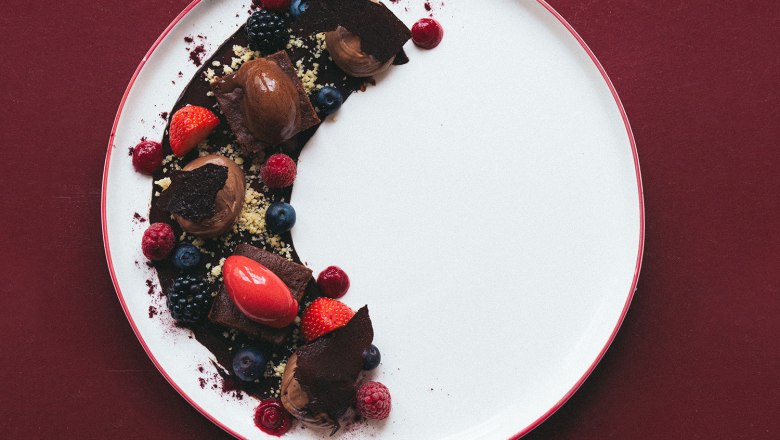 Ein Dessertteller mit Schokoladen-Mousse, Beeren und Schokoladenstücken auf rotem Hintergrund.