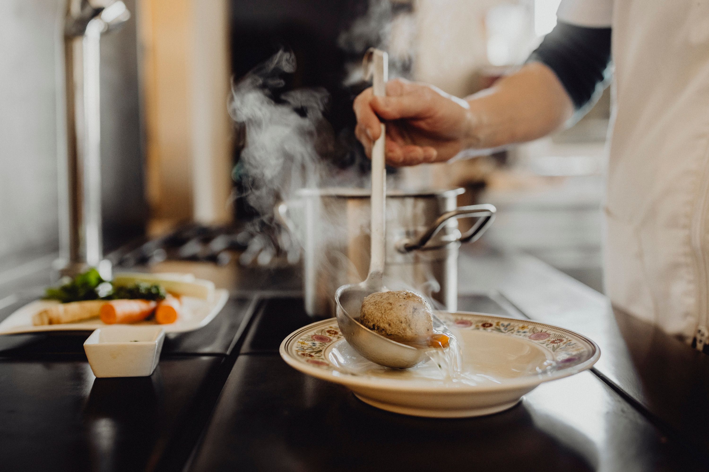 Eine Person schöpft Suppe mit einem Knödel aus einem Topf in einen Teller.