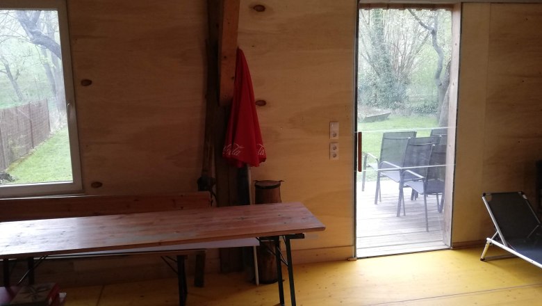 Innenansicht eines Raumes mit Holzw&auml;nden, Tisch, Liegestuhl und Blick auf Terrasse und Garten.