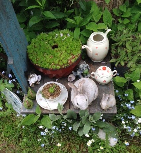 Gartenarrangement mit Teekanne, Tasse, Hasenfiguren und Sukkulenten.