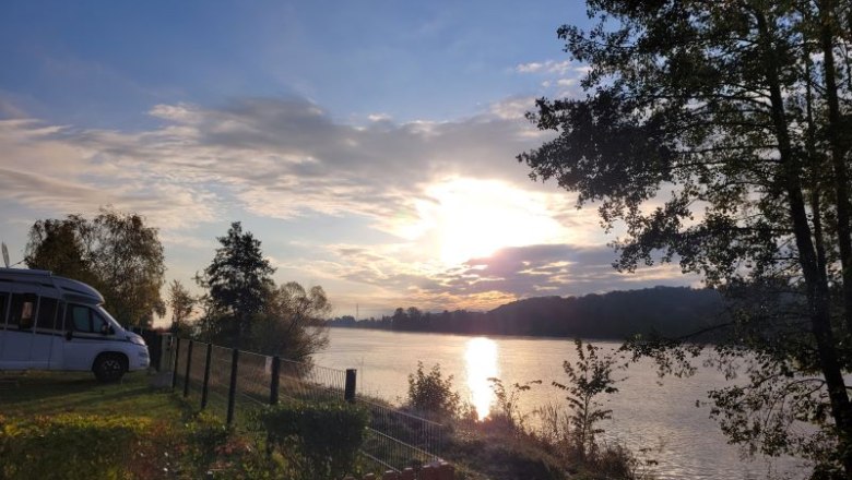 Sonnenuntergang &uuml;ber einem Fluss mit Wohnmobil und B&auml;umen im Vordergrund.