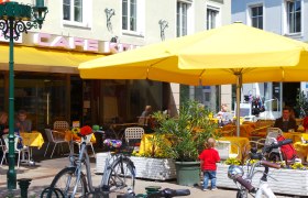 Außenbereich eines Cafés mit gelben Sonnenschirmen und Gästen an Tischen.