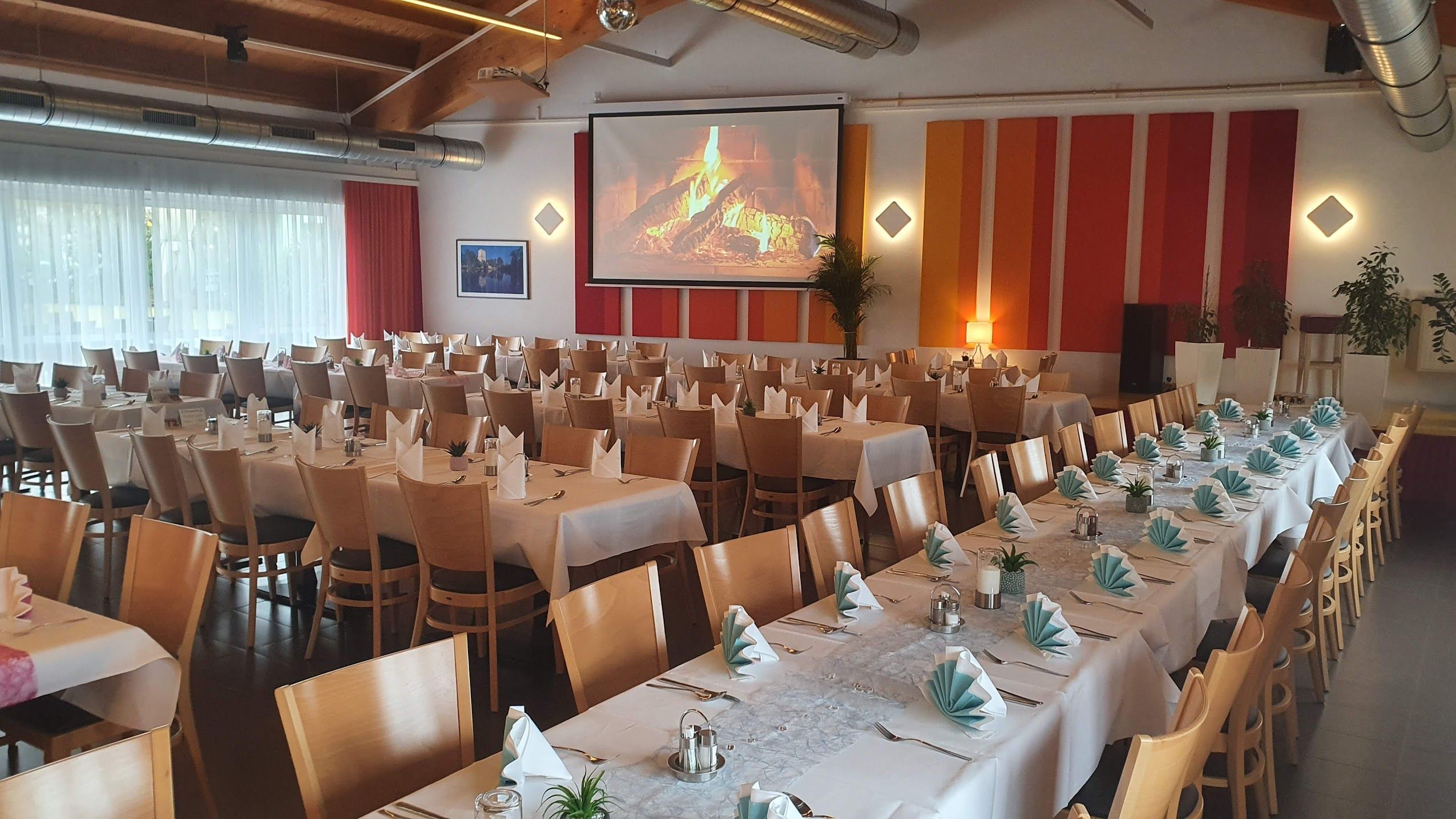 Ein festlich gedeckter Speisesaal mit langen Tischen, weißen Tischdecken und gefalteten Servietten. An der Wand ein Bildschirm mit Kaminfeuer.