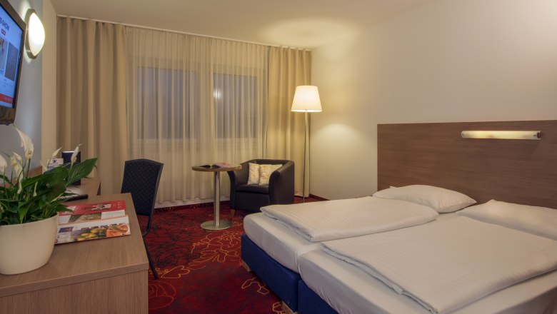 Doppelzimmer mit rotem Teppichboden, Schreibtisch und großem Doppelbett.