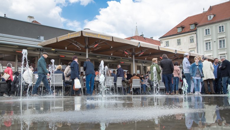 Menschen sitzen und stehen um einen Springbrunnen auf einem Platz mit Caf&eacute;.