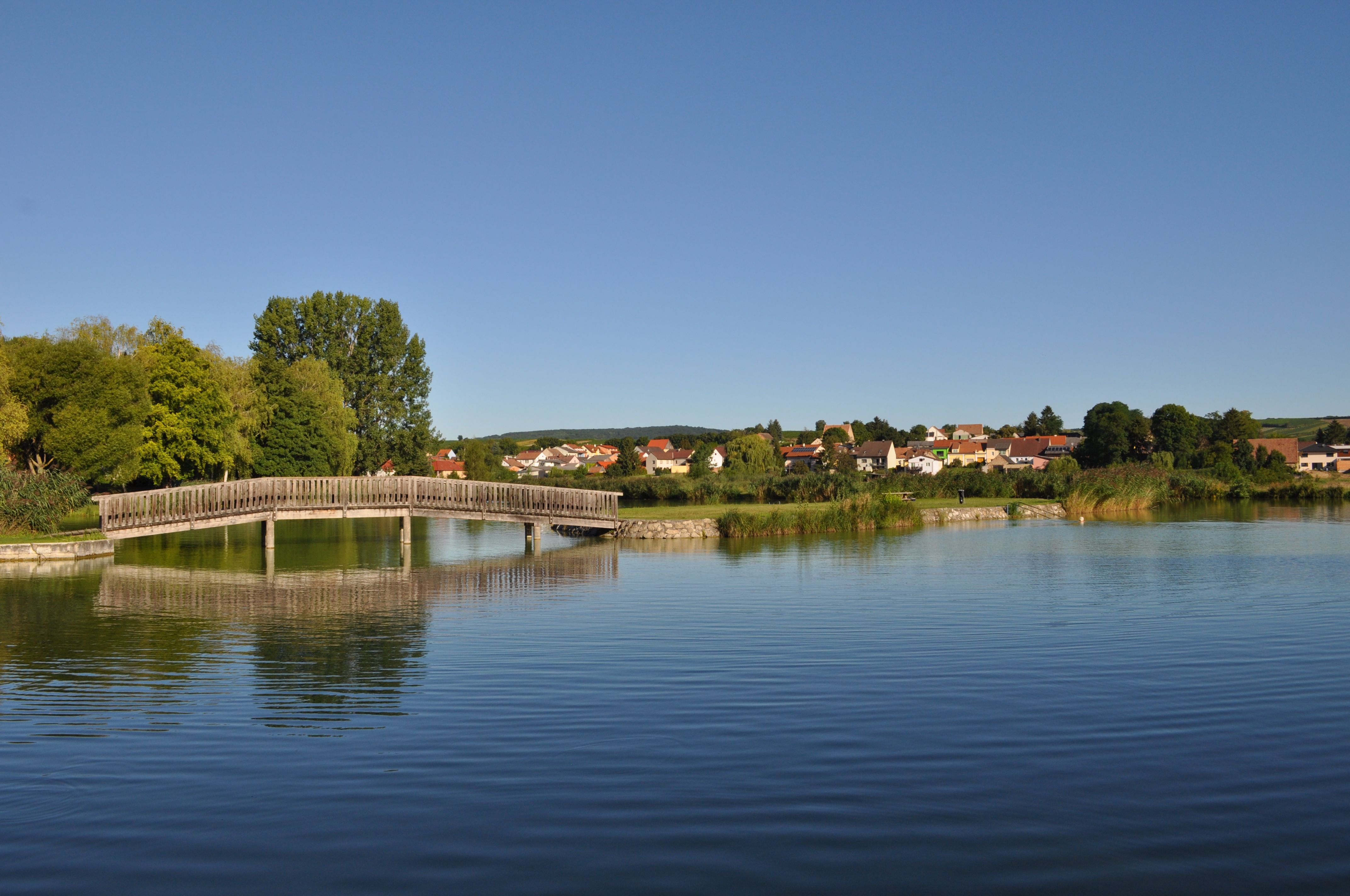 Holzbrücke über einen ruhigen Teich mit Dorf im Hintergrund.