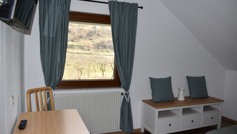 Innenansicht eines Zimmers mit Fenster, Tisch, Stuhl und Kommode.