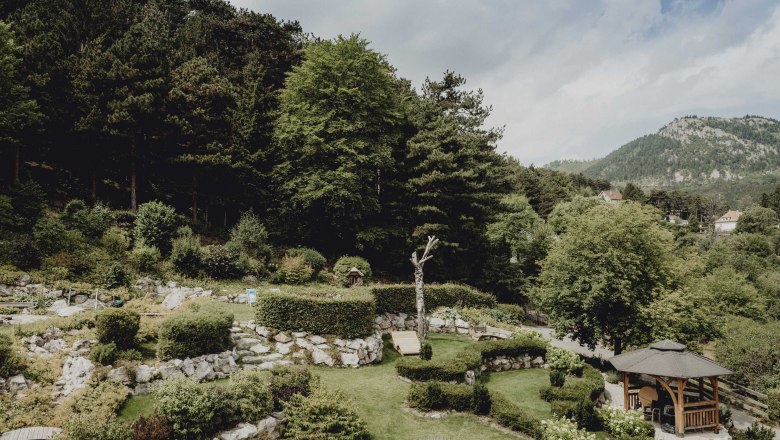 Ein Alpengarten mit B&auml;umen, Str&auml;uchern und einem Pavillon vor einer bewaldeten Berglandschaft.
