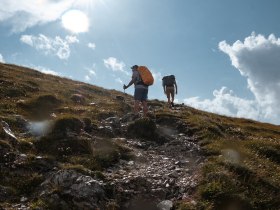 Die Wanderer erklimmen den sanften Hang, umgeben von &uuml;ppigem Gr&uuml;n und dem strahlenden Licht der Sonne. Die frische Bergluft und die atemberaubenden Ausblicke laden dazu ein, die Sch&ouml;nheit der Natur in vollen Z&uuml;gen zu genie&szlig;en.