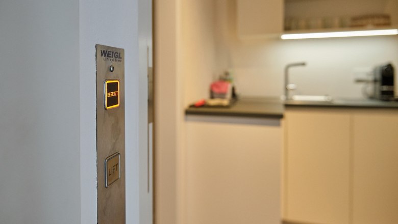 Ein Aufzugsknopf mit der Aufschrift 'Besetzt' leuchtet in einem modernen Wohnraum neben einer K&uuml;che.