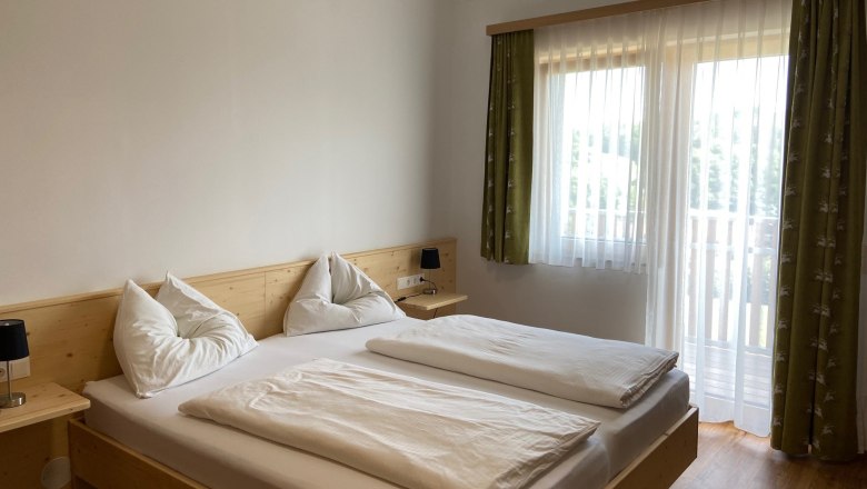 Ein helles Schlafzimmer mit einem Doppelbett, wei&szlig;en Bettdecken und Kissen. Ein Fenster mit gr&uuml;nen Vorh&auml;ngen l&auml;sst Tageslicht herein.