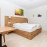 Modernes Schlafzimmer mit Holzmöbeln, Doppelbett, Tisch und Stuhl. Ein Spiegel und Bilder an der Wand, Tür führt zu einem Badezimmer.