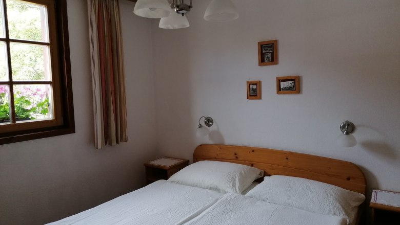 Gemütliches Doppelzimmer mit Holzmöbeln, einem Fenster mit Vorhängen und kleinen Bildern an der Wand.
