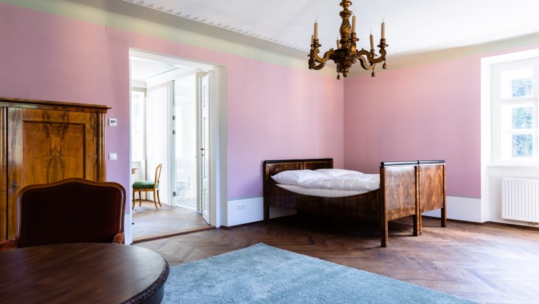 Ein elegantes Zimmer mit rosa W&auml;nden, einem Holzbett, einem Kronleuchter und einem Teppich auf Holzboden.