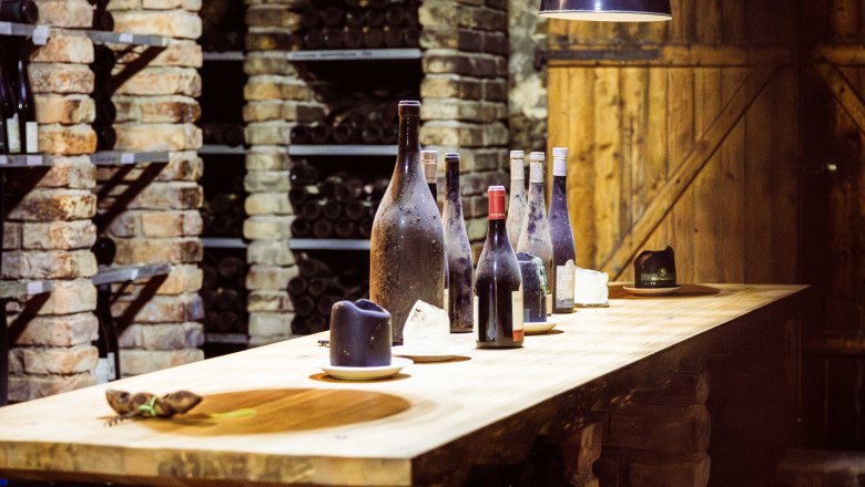 Ein Weinkeller mit Ziegelw&auml;nden, Regalen voller Weinflaschen und einem Holztisch mit mehreren Weinflaschen darauf.