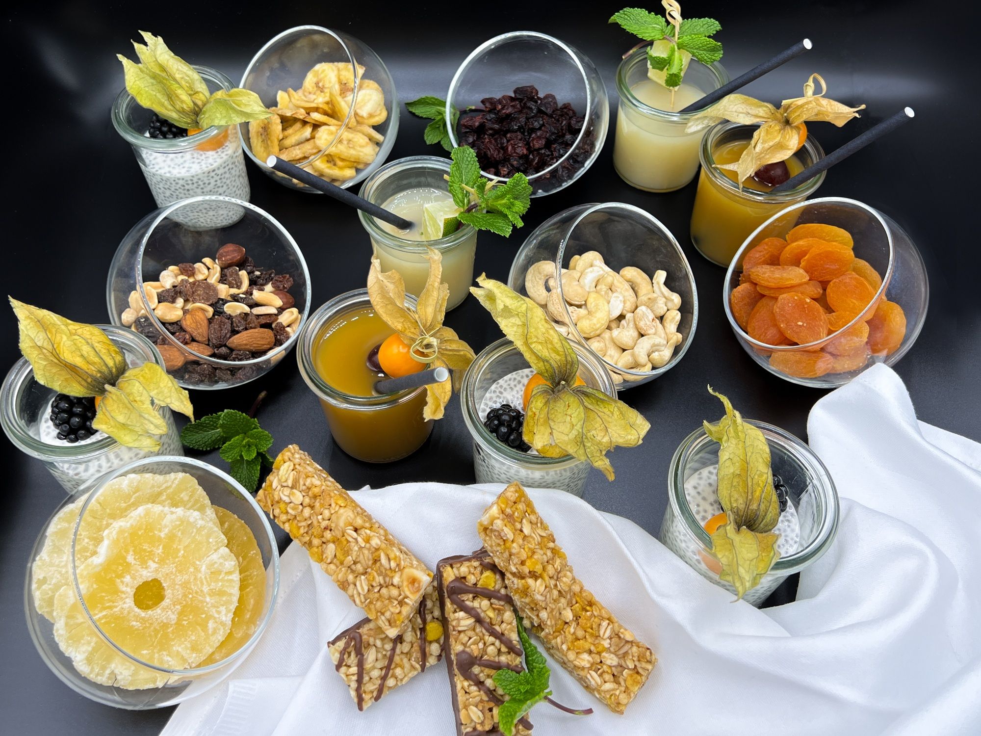 Eine Auswahl an Snacks und Getränken, darunter Nüsse, Trockenfrüchte, Chia-Pudding und Säfte, auf einem Tisch arrangiert.