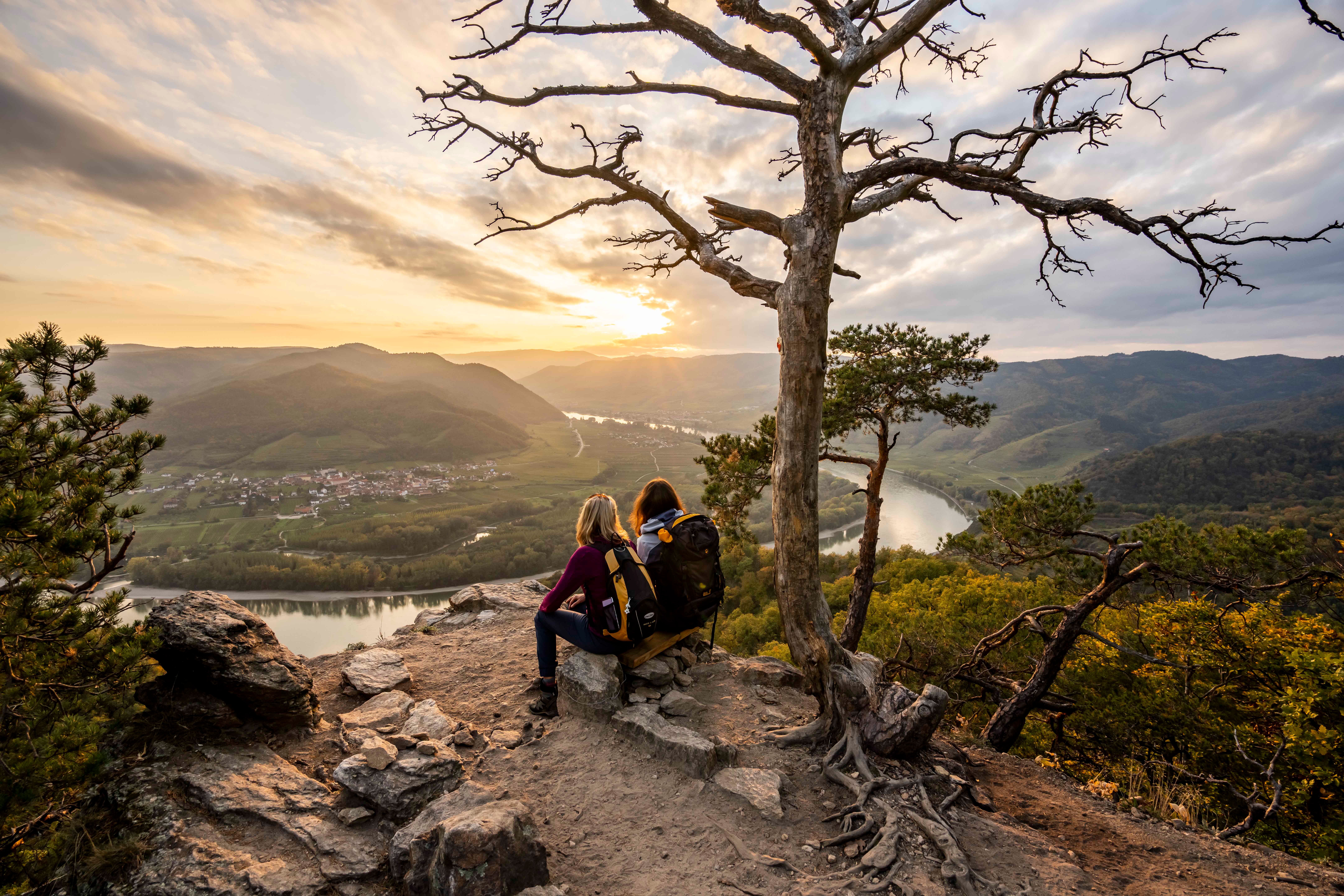 Zwei Personen sitzen auf einem Felsen und blicken auf eine Flusslandschaft bei Sonnenuntergang.