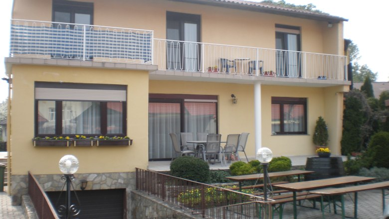 Zweistöckiges Haus mit Balkon und Terrasse, umgeben von Gartenmöbeln und Pflanzen.