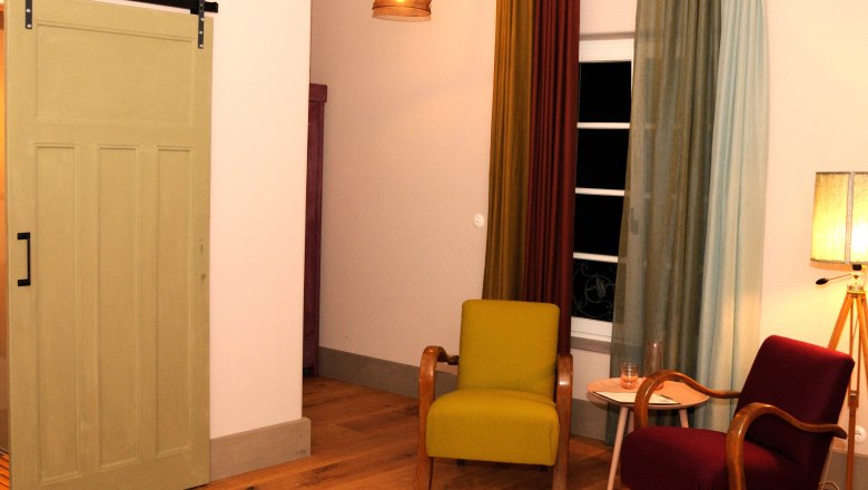 Ein gem&uuml;tliches Zimmer mit zwei Sesseln, einem kleinen Tisch und einer Lampe. Ein Schiebet&uuml;r und bunte Vorh&auml;nge sind ebenfalls zu sehen.