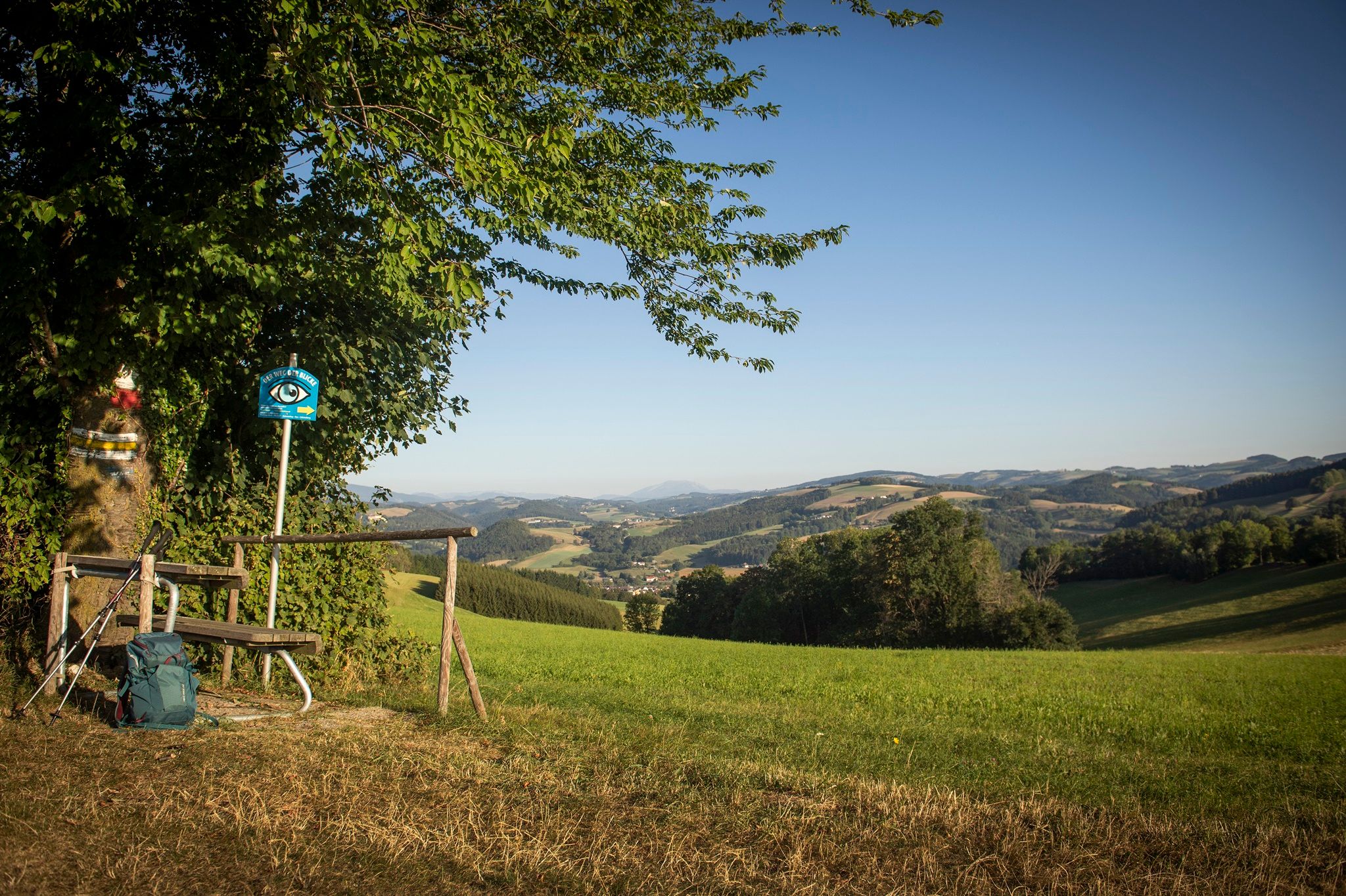 Landschaft mit Bank, Wanderstock und Rucksack im Vordergrund, Hügel und Berge im Hintergrund.