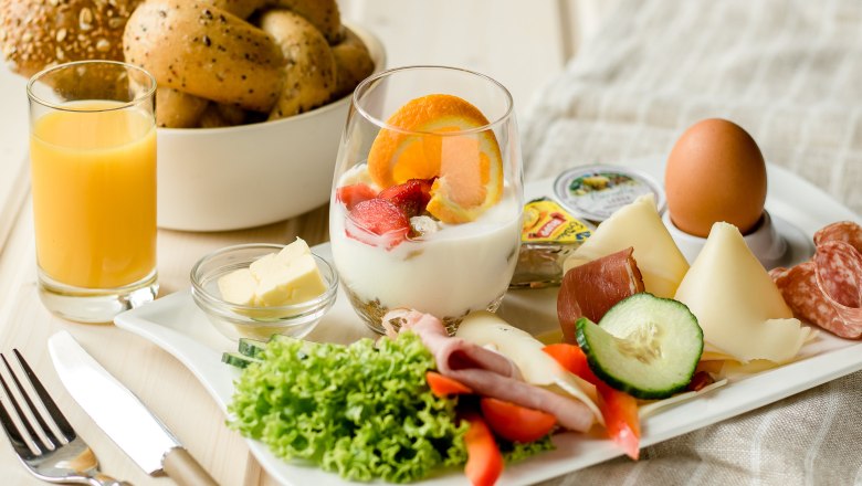 Ein Frühstückstablett mit Joghurt, Obst, Käse, Wurst, Ei, Salat, Butter und Orangensaft.