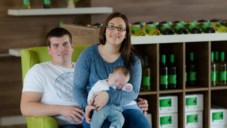 Familie Lust, © Astrid Bartl Ein Paar mit einem Baby sitzt auf einem grünen Sessel in einem Weingut.
