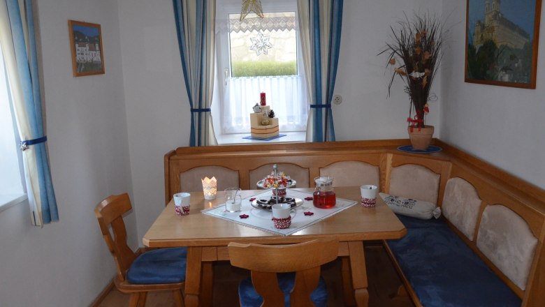 Gem&uuml;tliches Esszimmer mit Holztisch, Eckbank und blauen Polstern. Fenster mit blauen Vorh&auml;ngen und weihnachtlicher Dekoration.