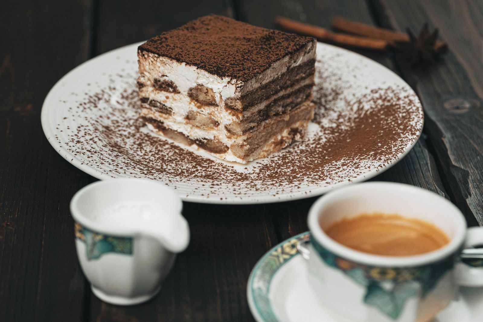 Ein Teller mit einem Stück Tiramisu, daneben eine Tasse Kaffee und ein Milchkännchen auf einem Holztisch.