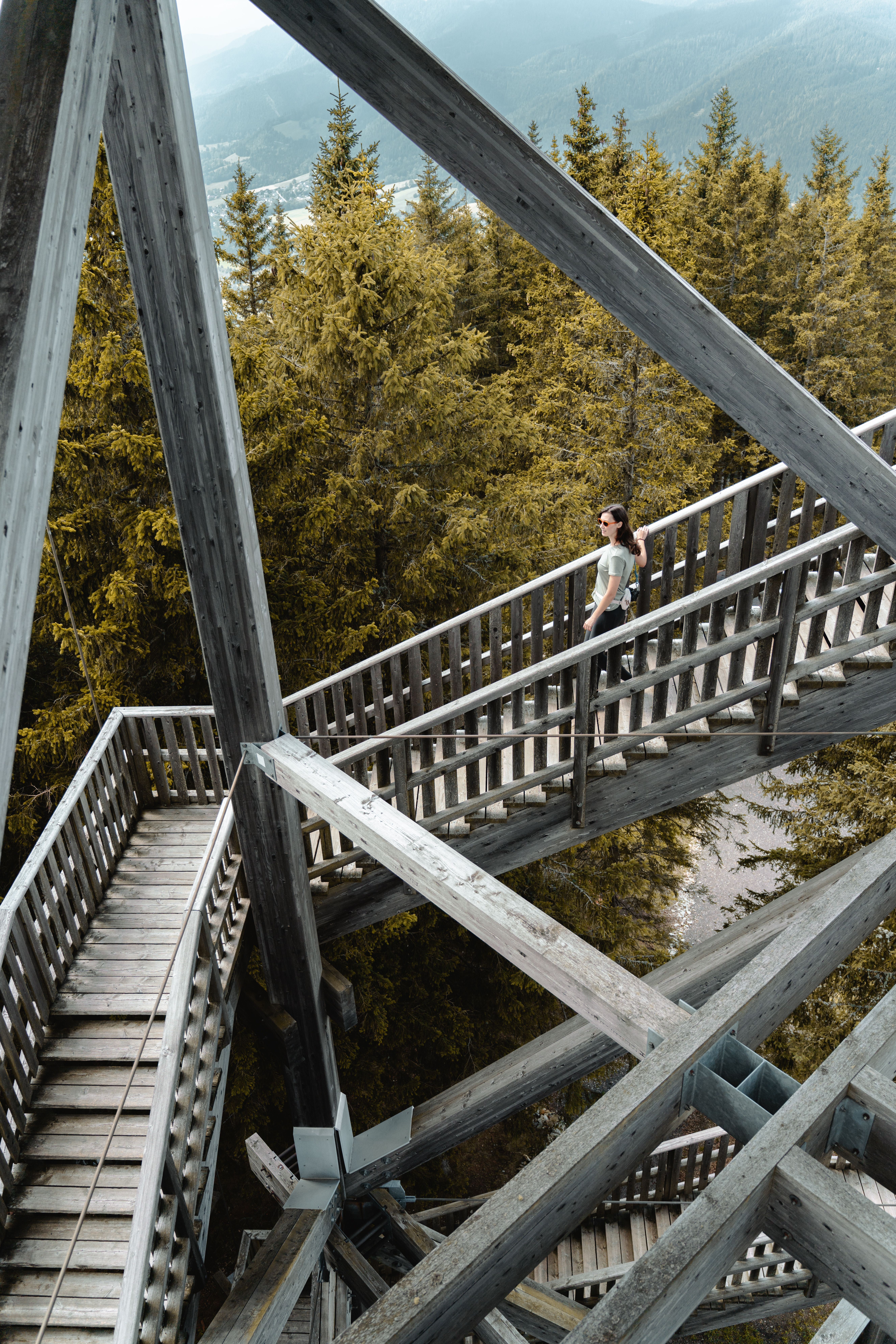 Zwei Wanderer gehen auf einem Weg bergauf, vorbei an einem Aussichtsturm und einem Gebäude, umgeben von Bäumen und einem klaren Himmel.