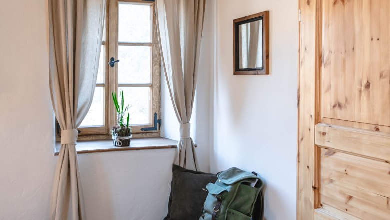 Ein kleiner Vorraum mit einem Fenster, Vorh&auml;ngen, einer Holzt&uuml;r und einer Sitzbank mit Rucksack und Decke.