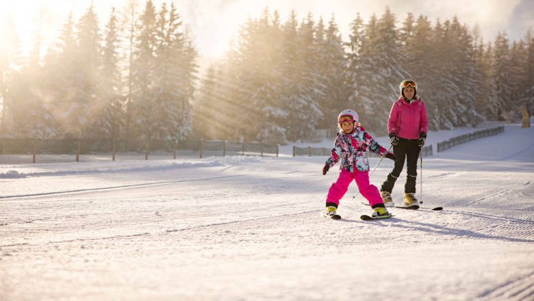 Kind und Erwachsener beim Skifahren im Schnee, umgeben von Bäumen und Sonnenstrahlen.