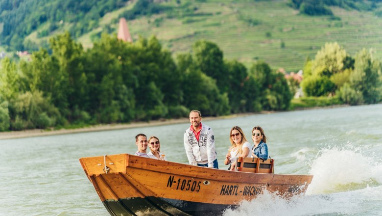Gruppe von Menschen auf einem Holzboot auf der Donau mit gr&uuml;ner Landschaft im Hintergrund.