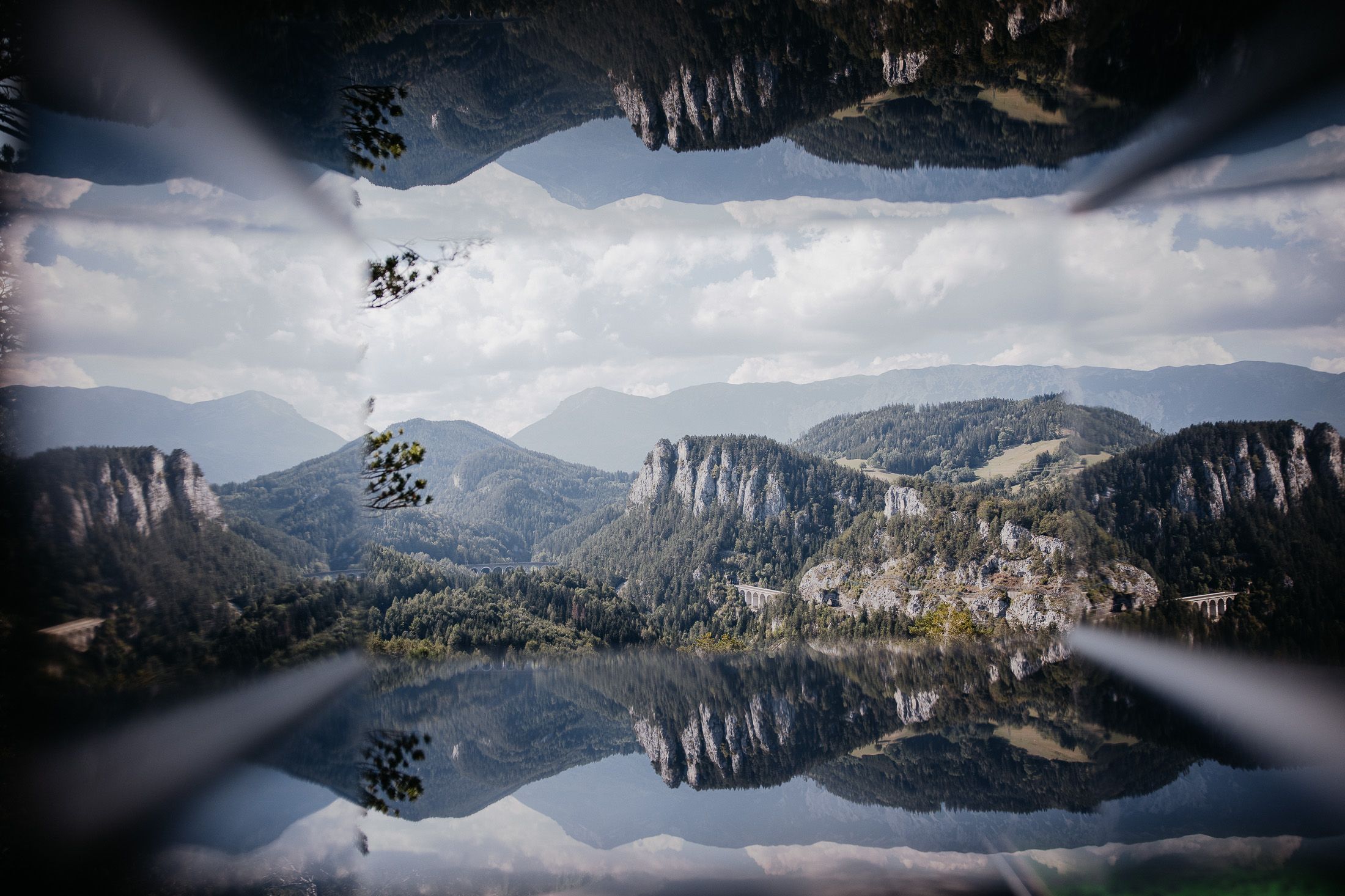 Spiegelbild einer Berglandschaft mit Viadukt und bewaldeten Hügeln unter blauem Himmel.