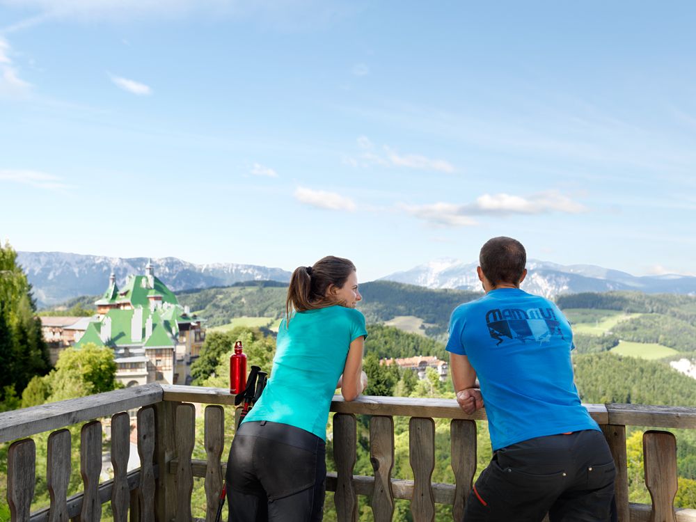 Zwei Personen stehen auf einem Balkon mit Blick auf das Südbahnhotel und die umliegenden Berge.