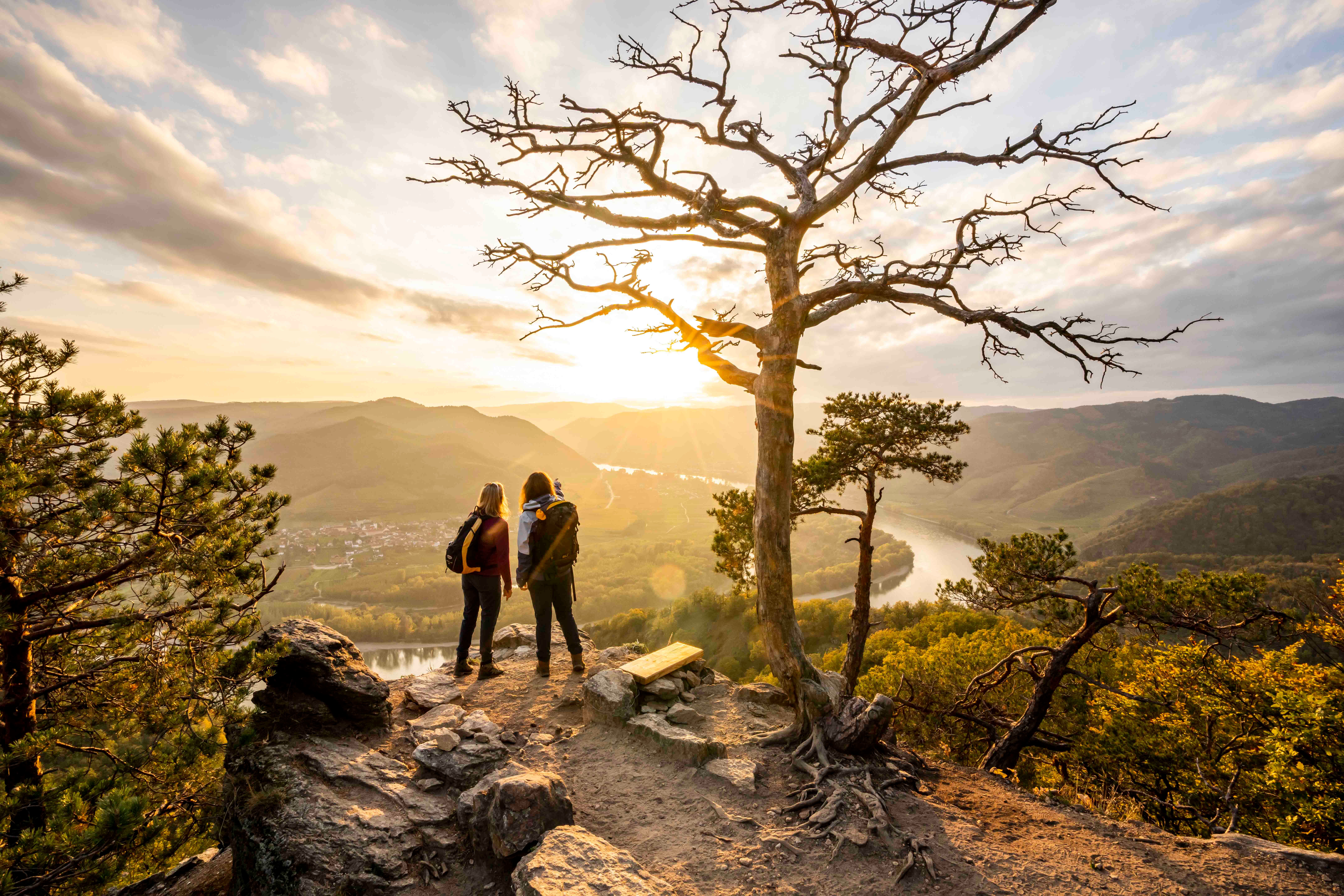 Zwei Personen stehen auf einem Felsen mit Blick auf eine Flusslandschaft bei Sonnenuntergang.