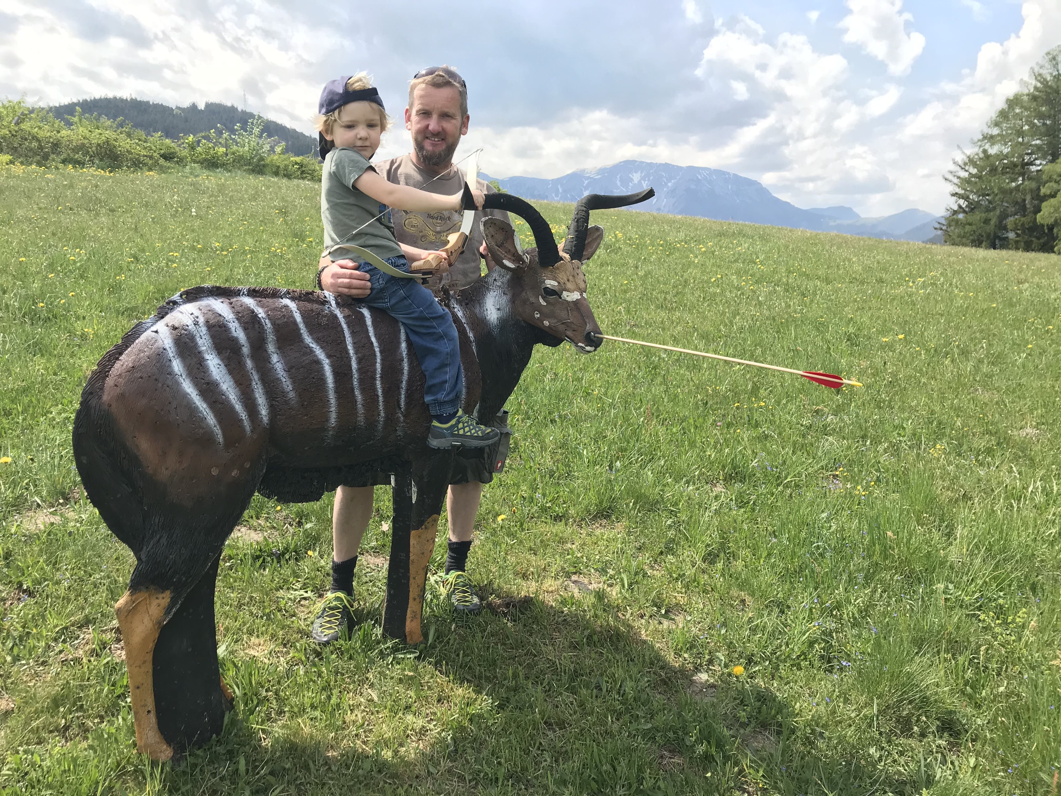 Ein Mann hält ein Kind auf einer 3D-Zielattrappe eines Antilopenartigen Tieres auf einer Wiese mit Bergen im Hintergrund.