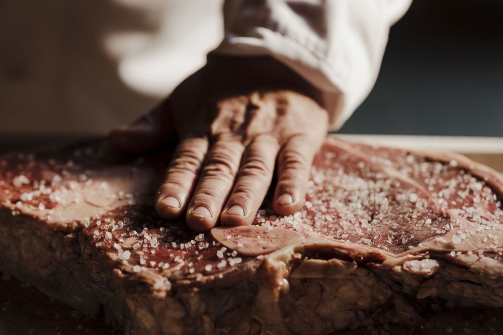 Nahaufnahme einer Hand, die grobes Salz auf ein großes Stück rohes Fleisch streut.